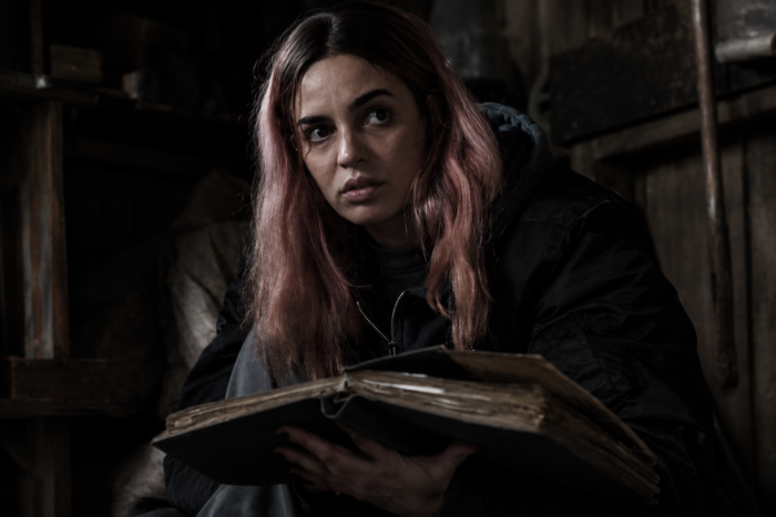 Evil Dead Burn Review: The Latest Entry Is a Bloody and Hilarious Good Time Woman with pink hair in a dim, wooden storage space holds an old, open book and looks off to the side.