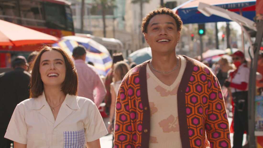 Gail Daughtry and the Celebrity Sex Pass Review: A Clever, Good Time with Wild Vibes and Offbeat Situations Two smiling adults walk through a sunny street market with colorful umbrellas and stalls in the background, enjoying the day.