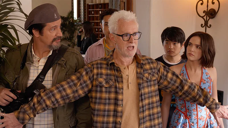 Gail Daughtry and the Celebrity Sex Pass Review: A Clever, Good Time with Wild Vibes and Offbeat Situations A gray-haired man with glasses stands center with arms outstretched, wearing a brown plaid shirt, while a small group watches him in a doorway area.