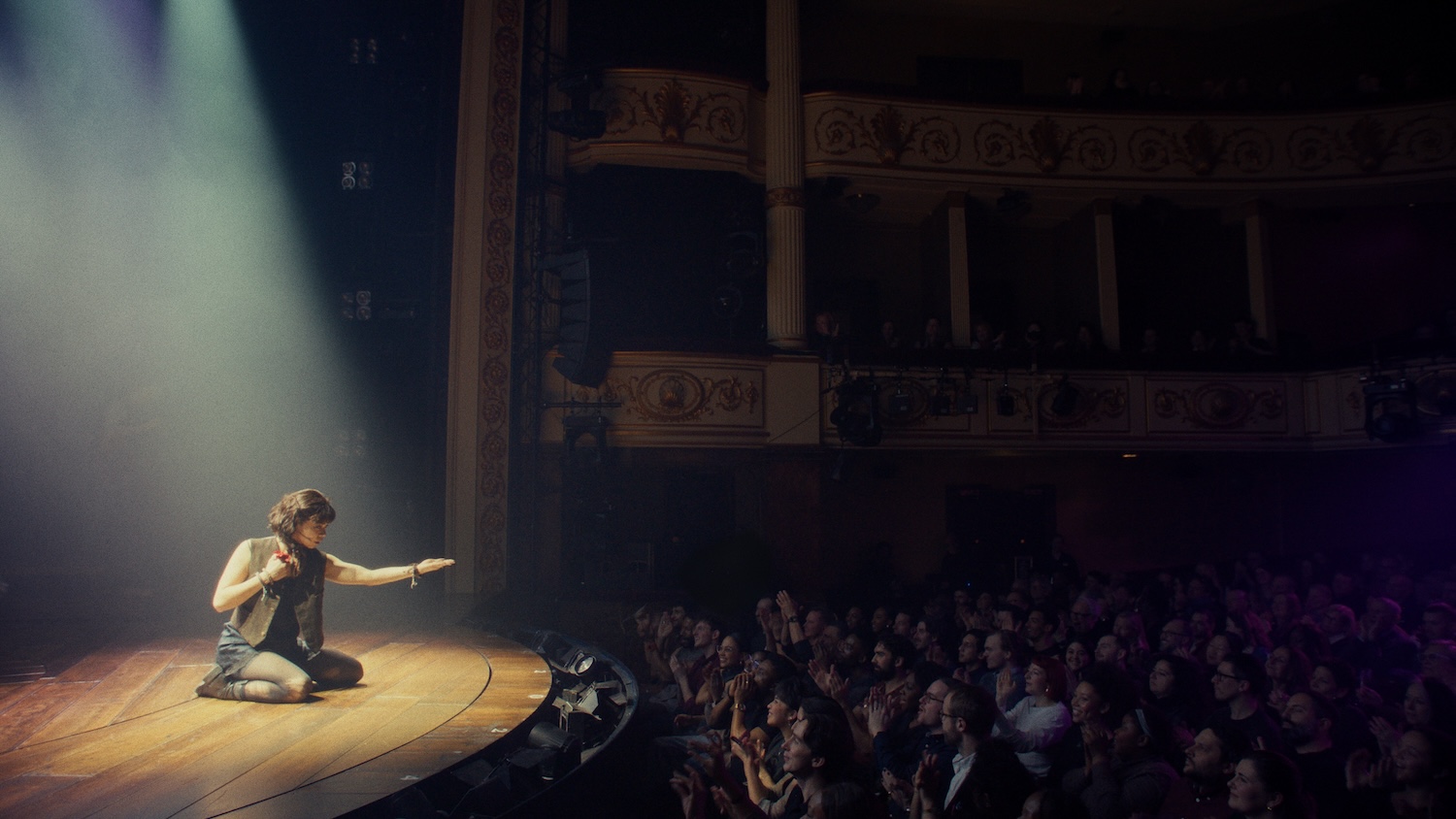 HADESTOWN_ THE MUSICAL Still 2 _ Courtesy of Crosswalk and LD Entertainmentjpeg - Eulalie Magazine Hadestown: The Musical Preview: Release Date, Cast, Plot, and More Performer kneels on a lit stage, reaching toward a cheering audience in a decorated historic theater setting.