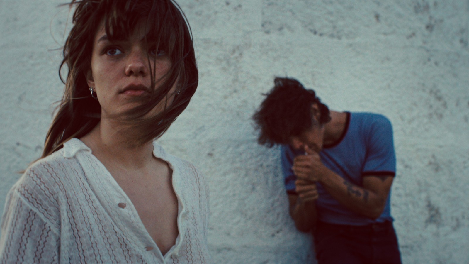 Romeria-2 - Eulalie Magazine Romería Review: A Layered and Fascinating Look at One Young Girl’s Family History Woman in a white blouse gazes at the camera as a man leans against a textured white wall in the background.