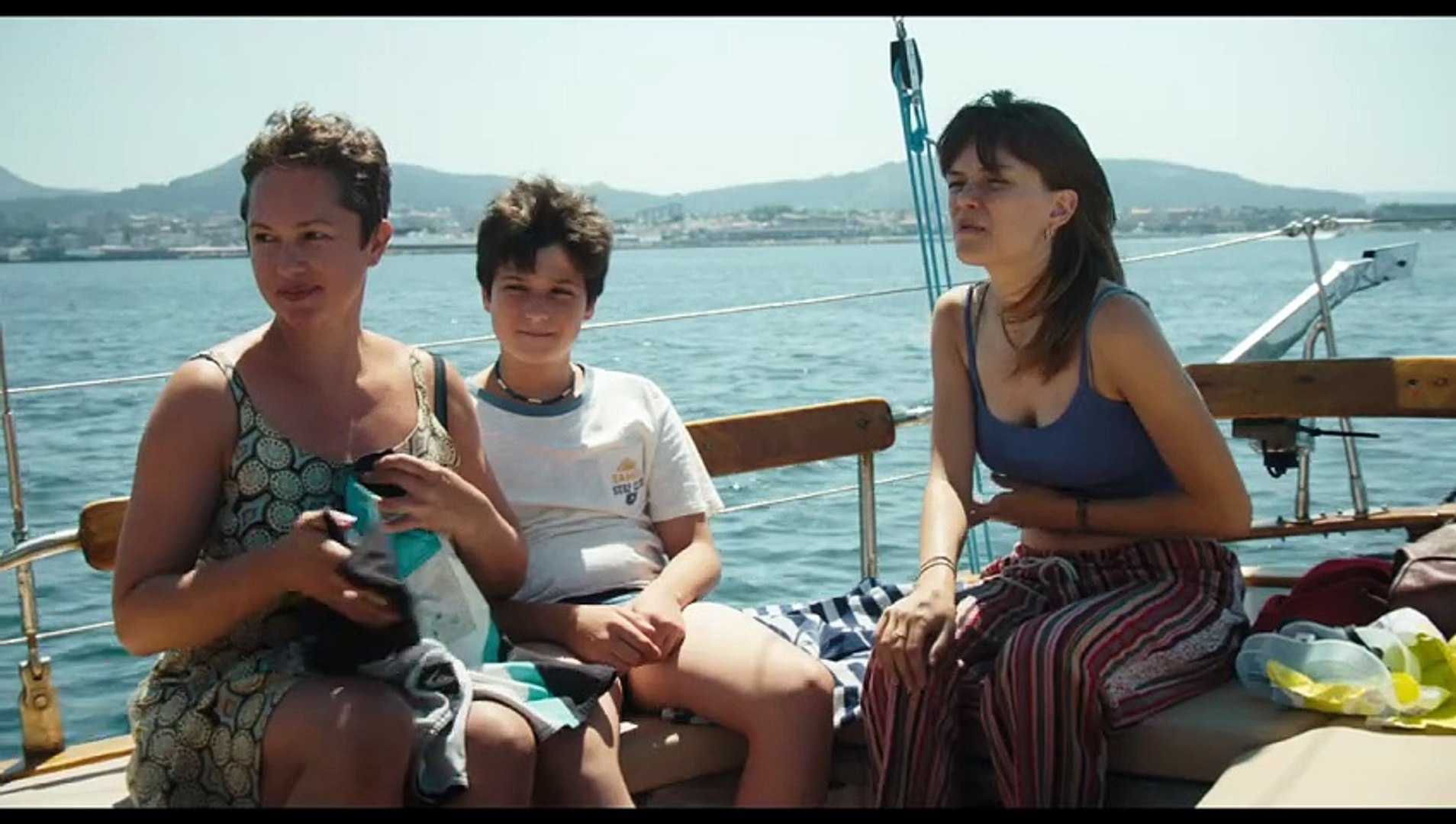 Romeria-3 - Eulalie Magazine Romería Review: A Layered and Fascinating Look at One Young Girl’s Family History Three people sit on a boat deck by the railing, with blue water and distant hills in the background.