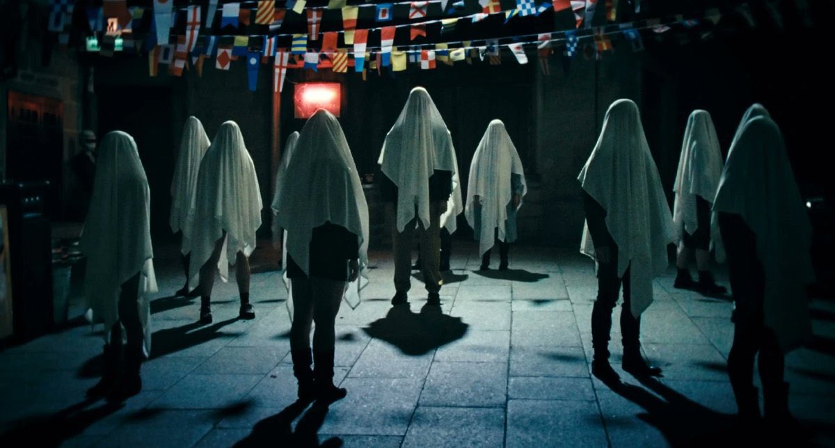 Romeria-4 - Eulalie Magazine Romería Review: A Layered and Fascinating Look at One Young Girl’s Family History People wearing white sheets over their heads stand in a circle in a dimly lit room with colorful flags overhead, creating a ghostly performance scene.