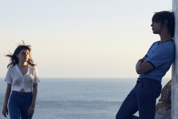 Romería Review: A Layered and Fascinating Look at One Young Girl’s Family History A woman in a white blouse and jeans stands by the sea with wind-blown hair as a man in a blue shirt leans against a white wall nearby.