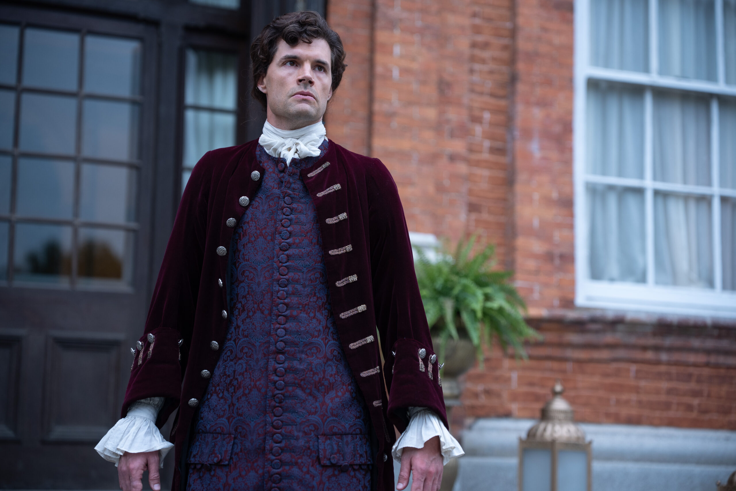 Young Washington Preview: Release Date, Cast, Plot, and More Man in 18th-century attire, burgundy velvet coat, white cravat, and blue patterned waistcoat stands outside a brick building.