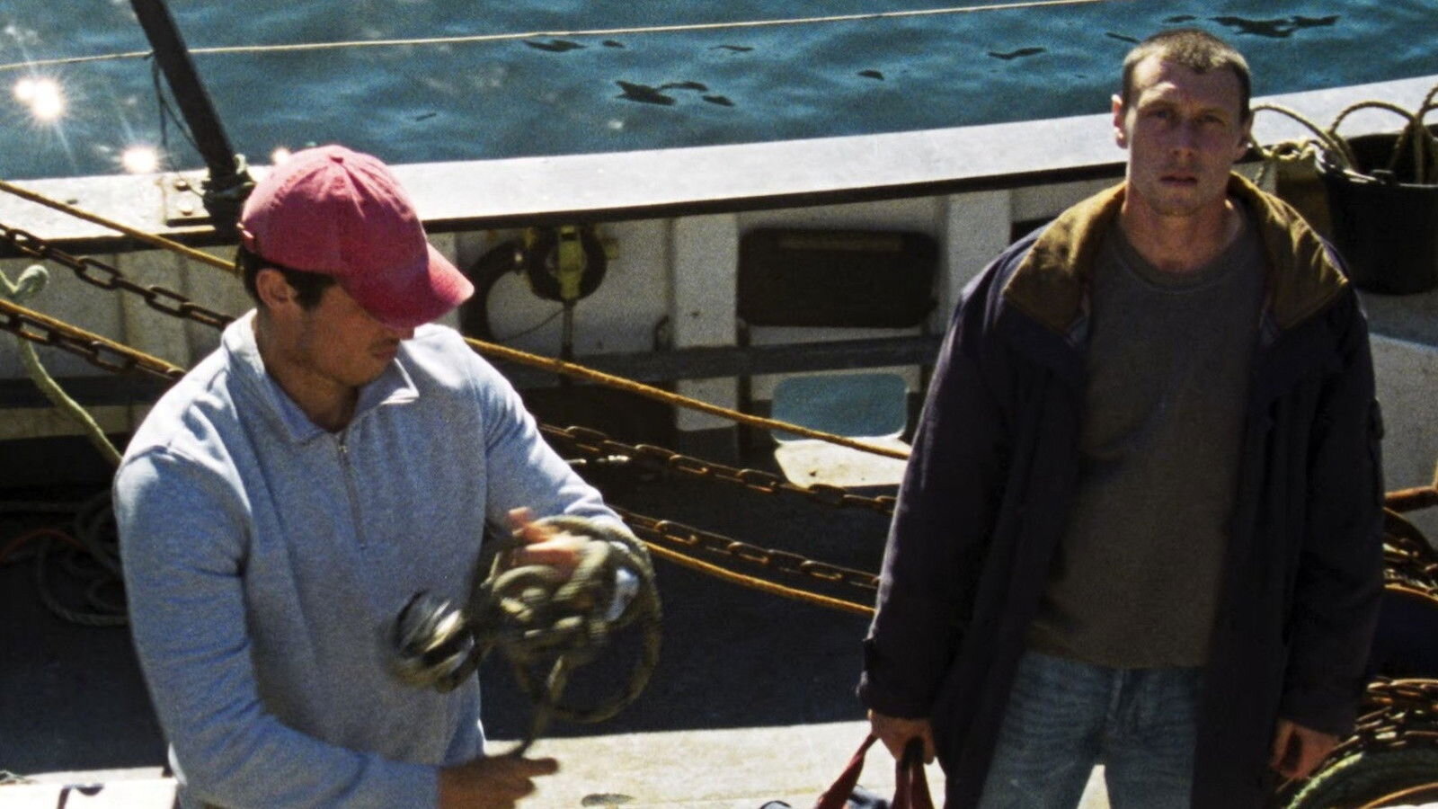 rose-of-nevada-3 - Eulalie Magazine Rose of Nevada Review: A Slow-Burn Movie That Sneaks Up on You and Hits Hard Two men on a fishing boat; one wearing a pink cap handles a fishing reel while the other man stands nearby in a dark jacket amid ropes and equipment by the water.