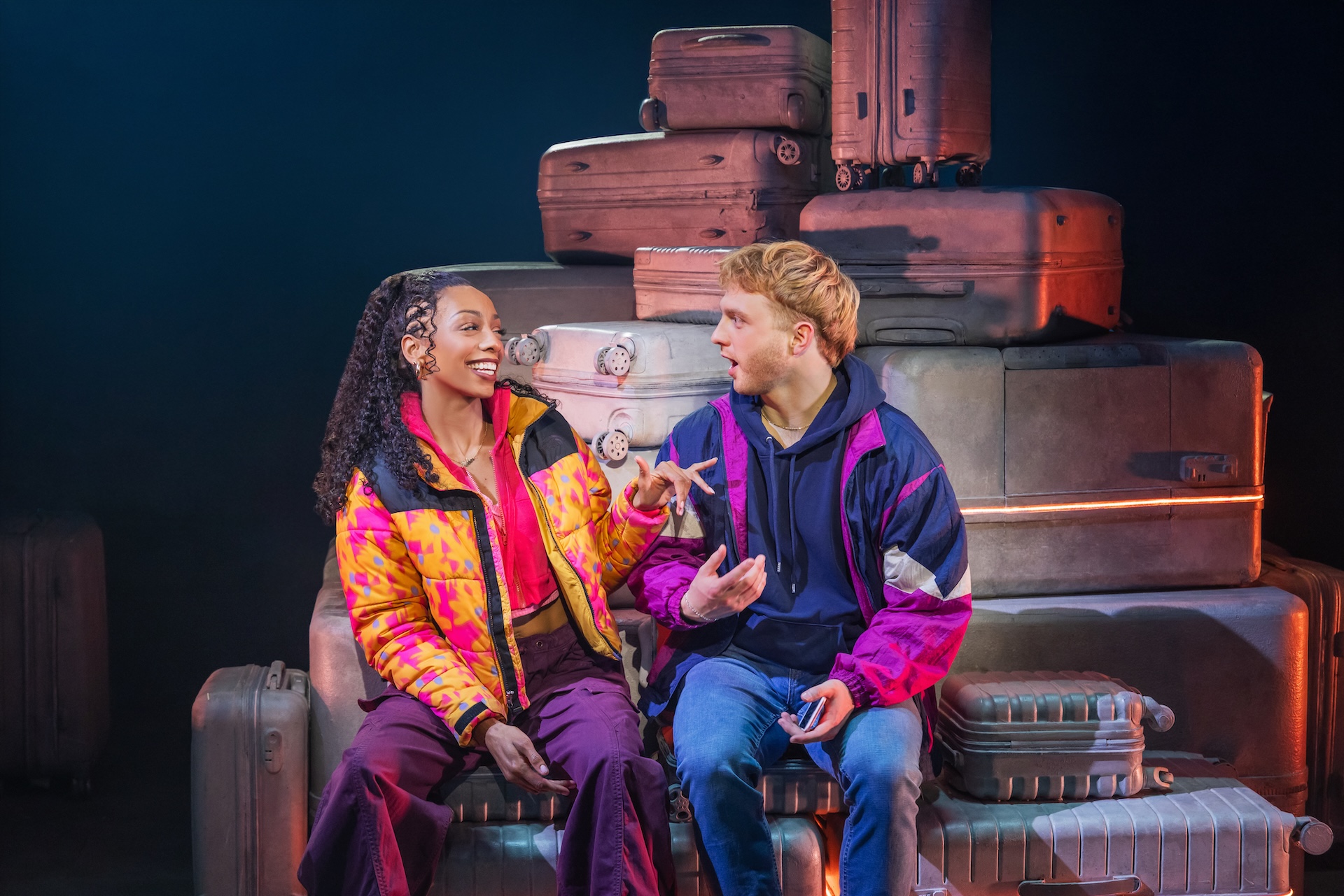 Two Strangers (Carry a Cake Across New York) Review: Relatable Situations and Memorable Characters Make for a Fun Musical Two young adults sit on stacked vintage suitcases, chatting and smiling backstage in colorful 90s outfits.