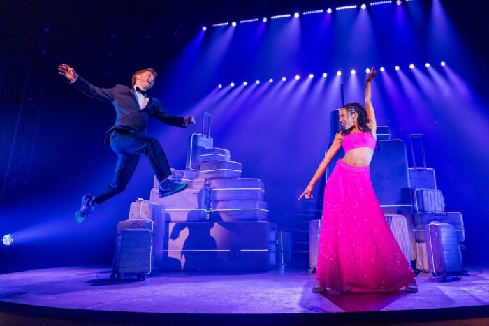 Two Strangers (Carry a Cake Across New York) Review: Relatable Situations and Memorable Characters Make for a Fun Musical Male dancer in a dark suit leaps across stacked suitcases on a purple-lit stage as a woman in a bright pink sparkly dress strikes a pose; dramatic lighting.