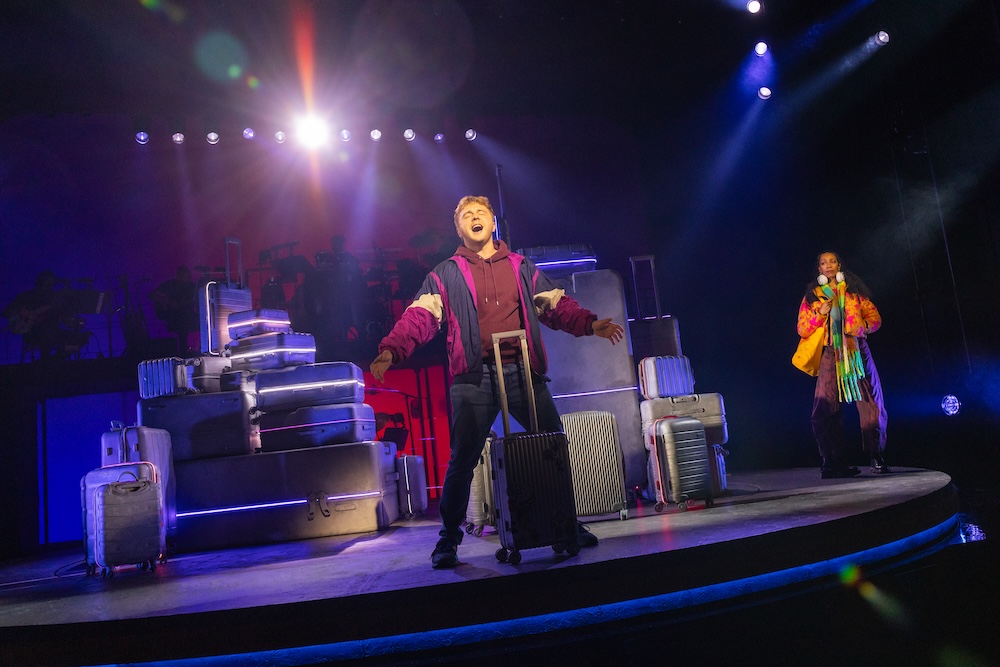 Two Strangers (Carry a Cake Across New York) Review: Relatable Situations and Memorable Characters Make for a Fun Musical Two vocalists perform on a circular stage with stacked suitcases as props and vivid stage lighting in the background.