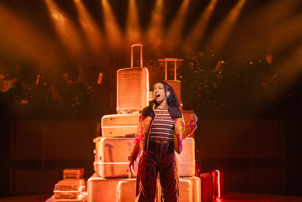 Two Strangers (Carry a Cake Across New York) Review: Relatable Situations and Memorable Characters Make for a Fun Musical Singer performs on stage beside stacked suitcases under warm orange lights, wearing a striped top and colorful jacket, mid-song pose.