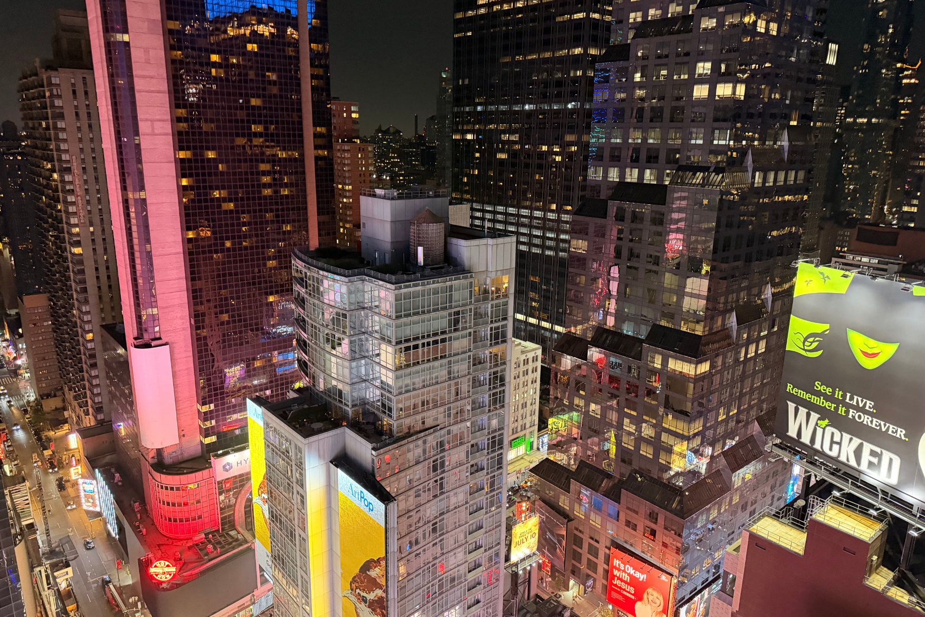 Times Square NYC - Eulalie Magazine First Time in New York City? Here’s Everything You Need to Know Before You Go! Nighttime aerial view of Times Square: tall glass buildings lit with colorful neon signs and large illuminated billboards filling the streets below, including a bright yellow Wicked ad on the right.