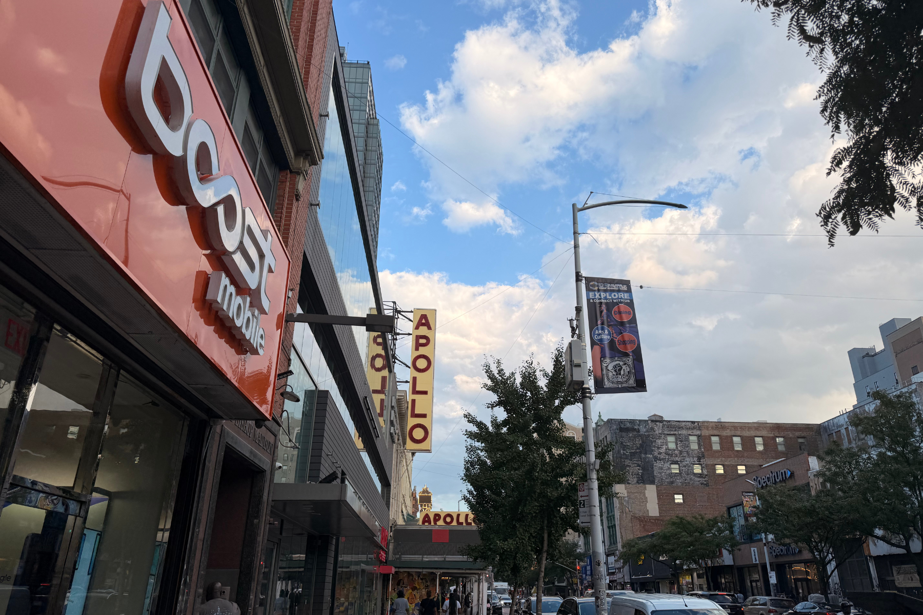Apollo Theater NYC - Eulalie Magazine First Time in New York City? Here’s Everything You Need to Know Before You Go! City street scene with a bold red storefront sign reading'ACS' or 'cos' on the left and an Apollo theater marquee across the street under a blue sky with clouds.