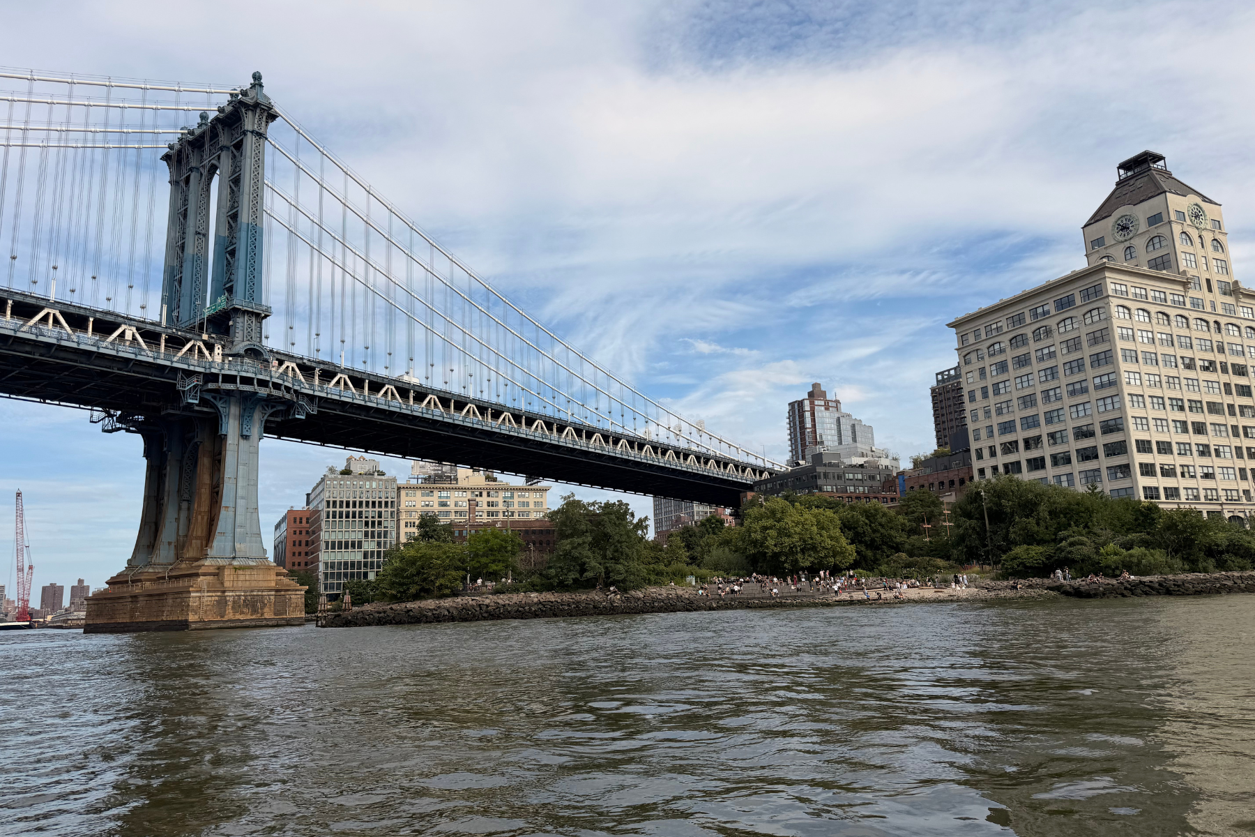 The Manhattan Bridge NYC - Eulalie Magazine First Time in New York City? Here’s Everything You Need to Know Before You Go! Suspension bridge over a river with tall towers, blue sky, and a cityscape in the background, with people along the shoreline.