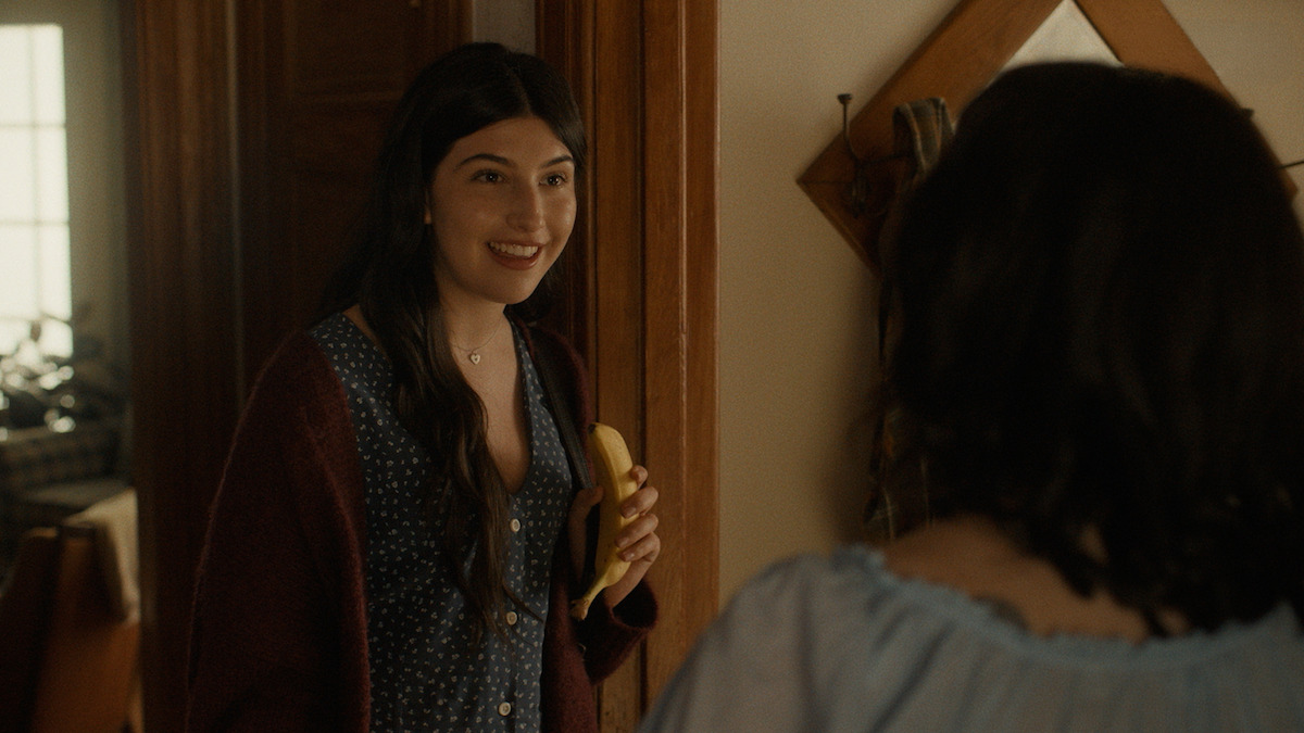 Dont-Say-Good-Luck-4 - Eulalie Magazine Don’t Say Good Luck Review: Sunny Sandler is Solid Gold in This Slice of Life Drama Smiling woman in a doorway holds a banana and talks to someone offscreen in a home interior.