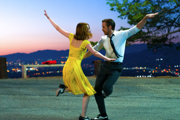 Couple dancing outdoors at dusk beside a cityscape, woman in yellow dress and man in white shirt and tie with arms raised in a dance pose
