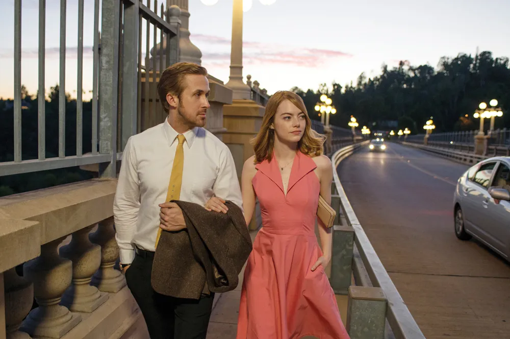 La-La-Land-2 - Eulalie Magazine La La Land 10th Anniversary Review: Romance Movie Will Still Leave Audiences on Cloud Nine Man and woman walk together on a lit bridge at sunset; the man wears a white shirt and yellow tie, holding a brown coat, while the woman wears a pink sleeveless dress and carries a beige clutch.