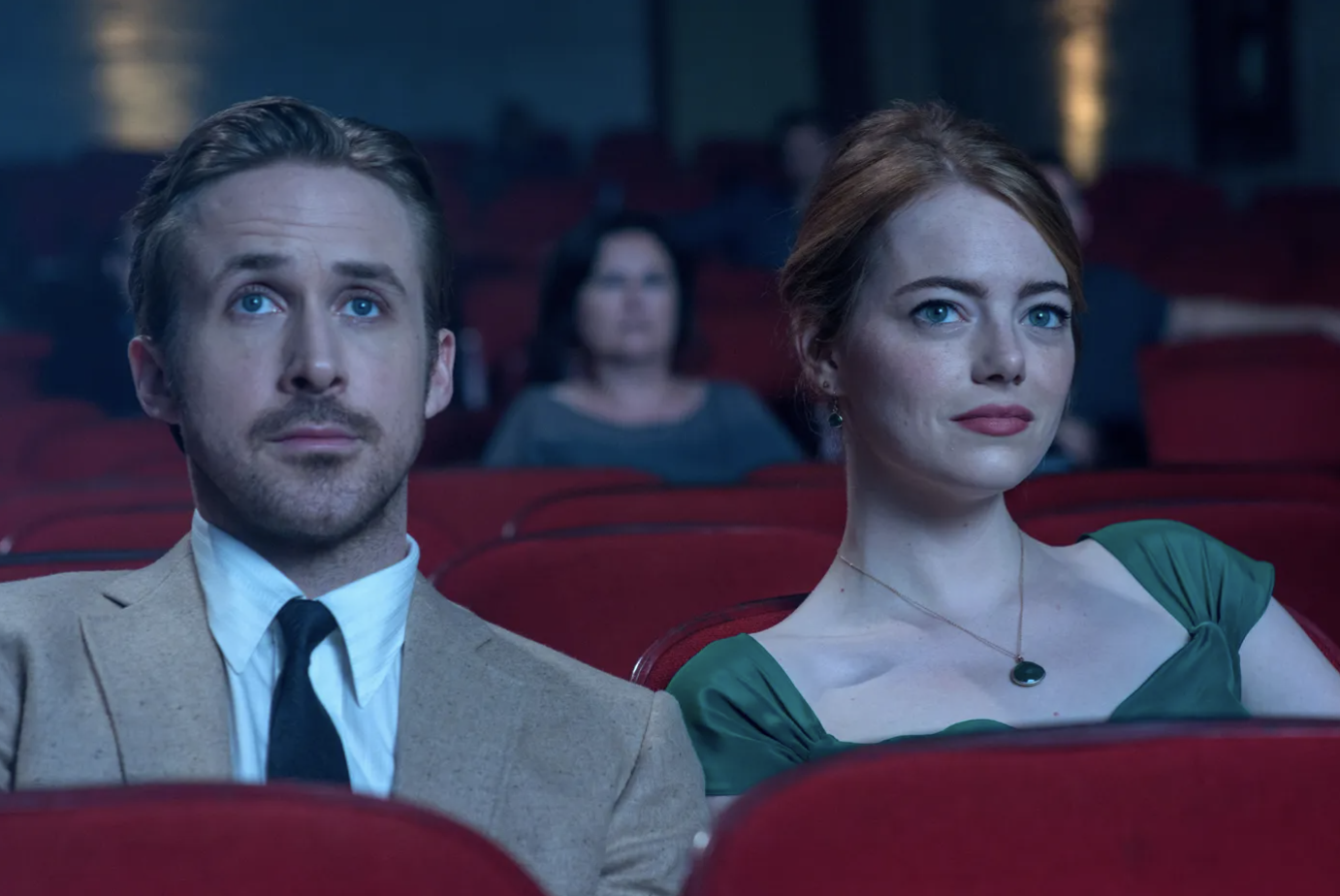 La-La-Land-3 - Eulalie Magazine La La Land 10th Anniversary Review: Romance Movie Will Still Leave Audiences on Cloud Nine A man in a beige suit and a woman in a green dress sit in a dim movie theater, looking toward the screen.