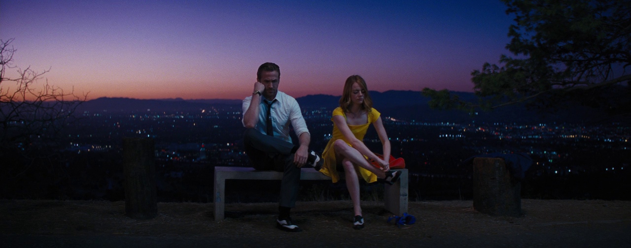 La-La-Land-4 - Eulalie Magazine La La Land 10th Anniversary Review: Romance Movie Will Still Leave Audiences on Cloud Nine Man in a white shirt and tie and a woman in a yellow dress sit on a bench atop a hill at dusk, city lights below.
