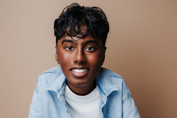 Actor Nylan Parthipan smiles for a headshot while sitting in front of a light brown background.
